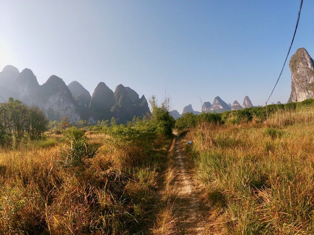 A dirt path surrounded by dry grass and with some mountains in the distance, taken while hiking from Xingping to Shawan.