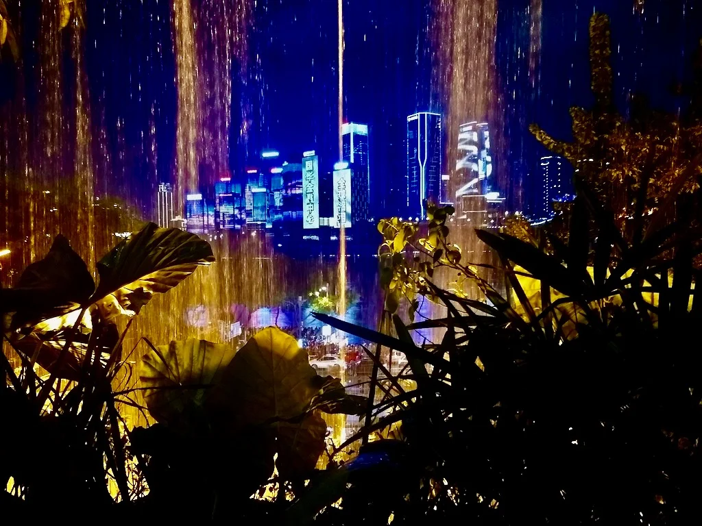 Views of lit-up buildings as water lightly cascades down in front of the camera. Located during travels to Chongqing, China, downtown