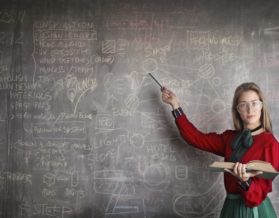 A teacher in a red sweater and holding a book with one hand points to a blackboard filled with words and numbers.