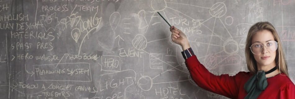 A teacher in a red sweater and holding a book with one hand points to a blackboard filled with words and numbers.
