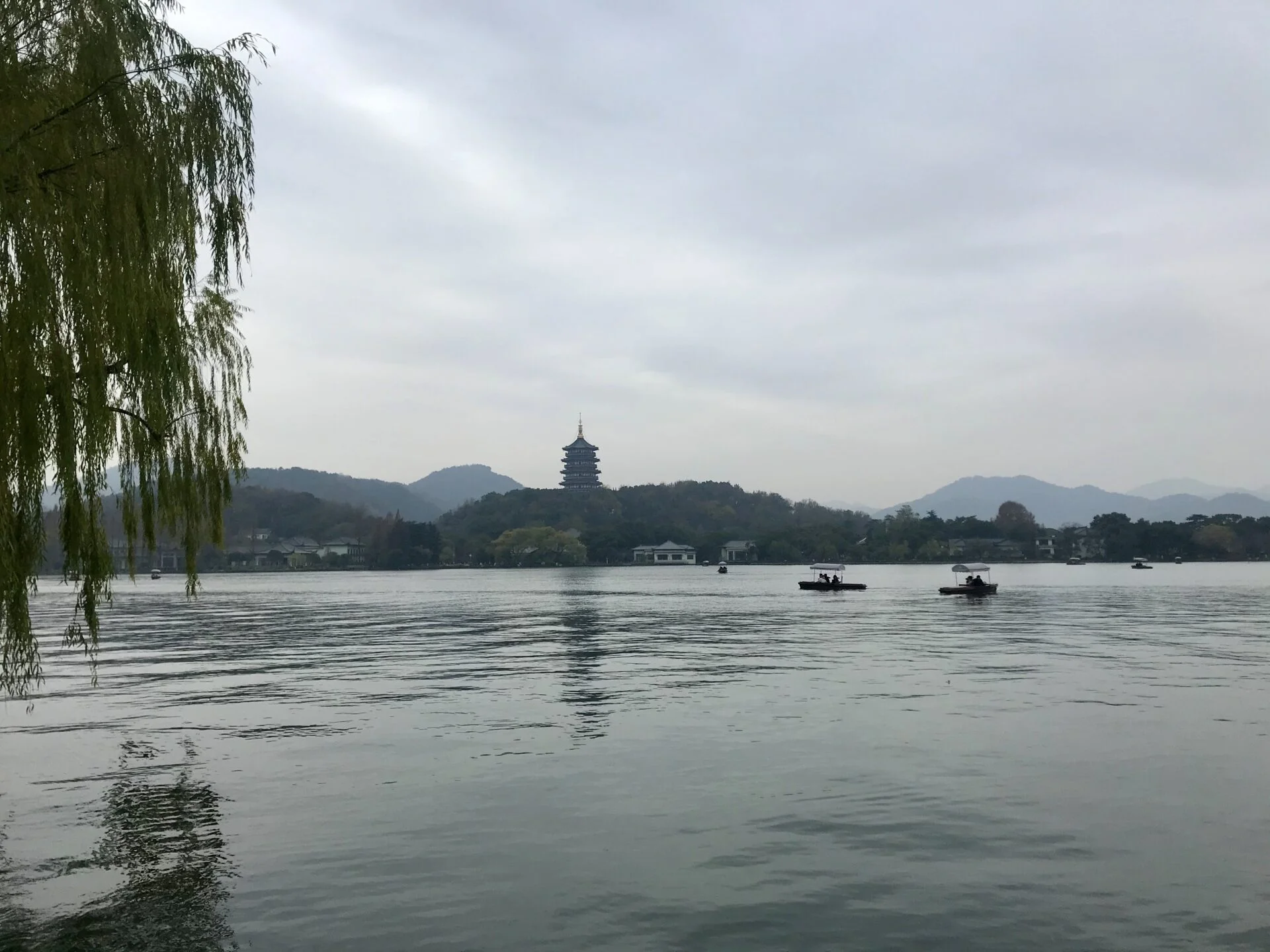 Forced to wrong size: 451x338, natural is 1920x1440! A calm blue lake in front. A green hanging plant on the left side. A Chinese template in the distance behind rolling hills. Two boats on the lake.