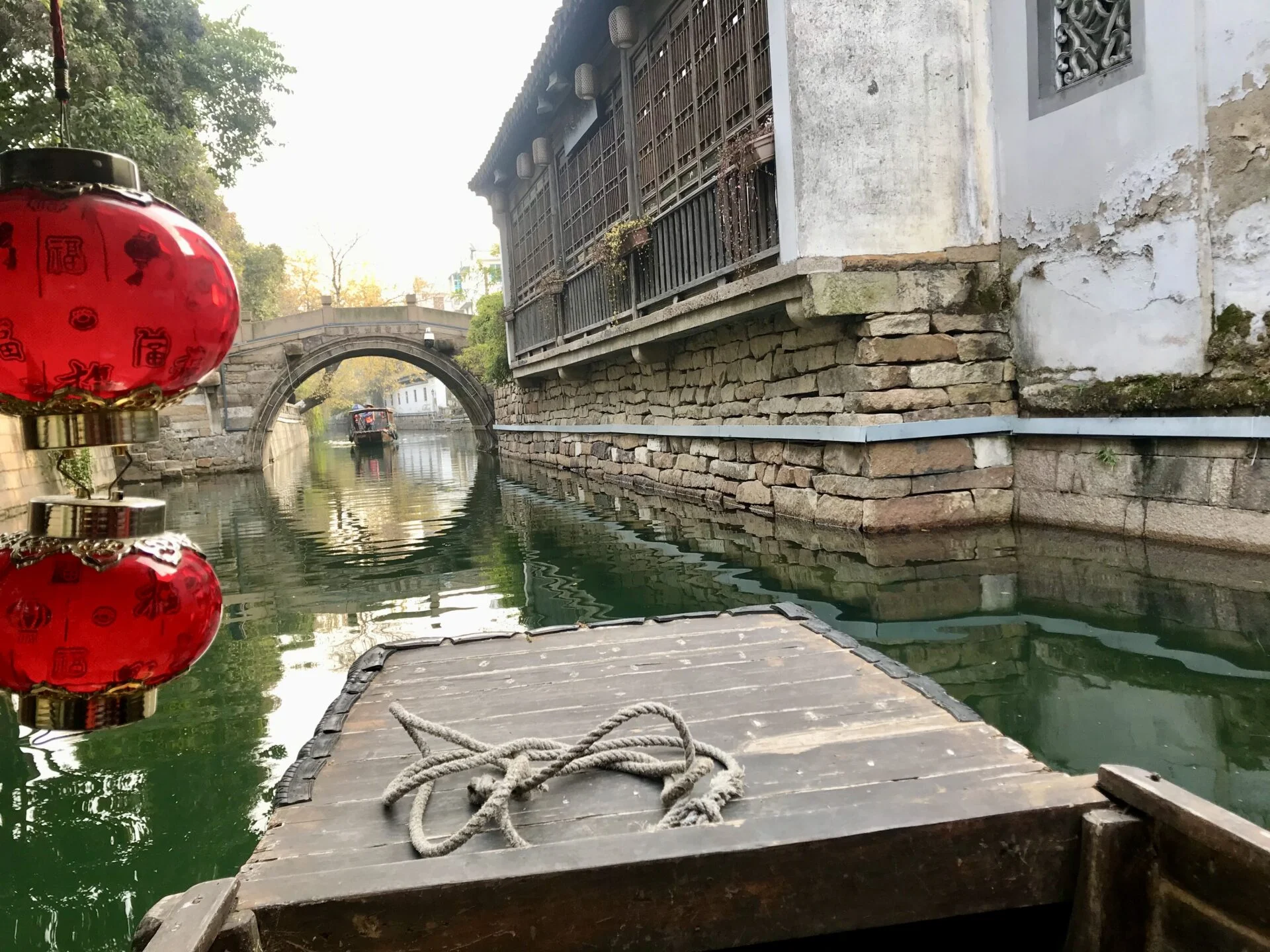 Forced to wrong size: 451x338, natural is 1920x1440! A wooden boat with a white rope on it. In a green-blue colored canal. A circular opening with a bridge ahead, and a large building on stones to the right. 2 red lanterns half-cut off on the left side.
